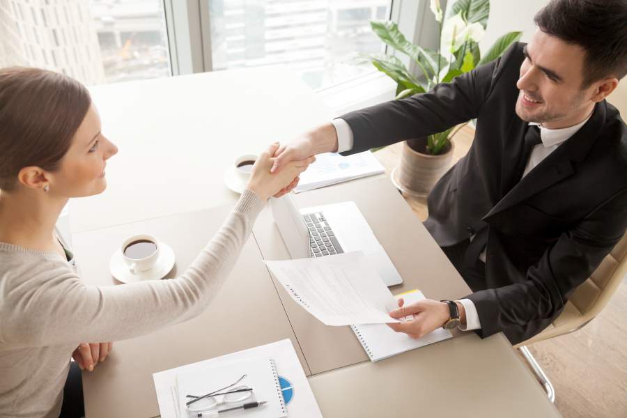 Geschäftsfrau und Geschäftsmann sitzen am Tisch mit Handshake, vor ihnen liegen Unterlagen in einem hellen Besprechungszimmer, sie lächeln