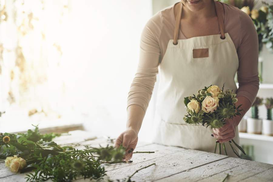 Floristin mit weißer Schürze macht einen frischen Blumenstrauß mit weißen Rosen in einem freundlichen, hellen Blumenladen