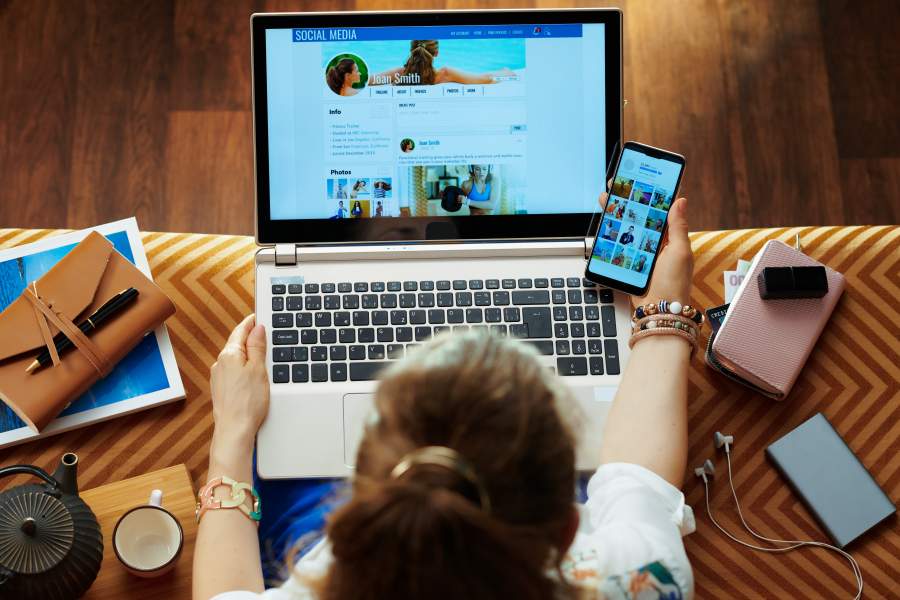 Frau sitzt mit Laptop auf dem Schoß und einem Smartphone in der rechten Hand auf einem Sofa und checkt ihre Social Media Accounts.