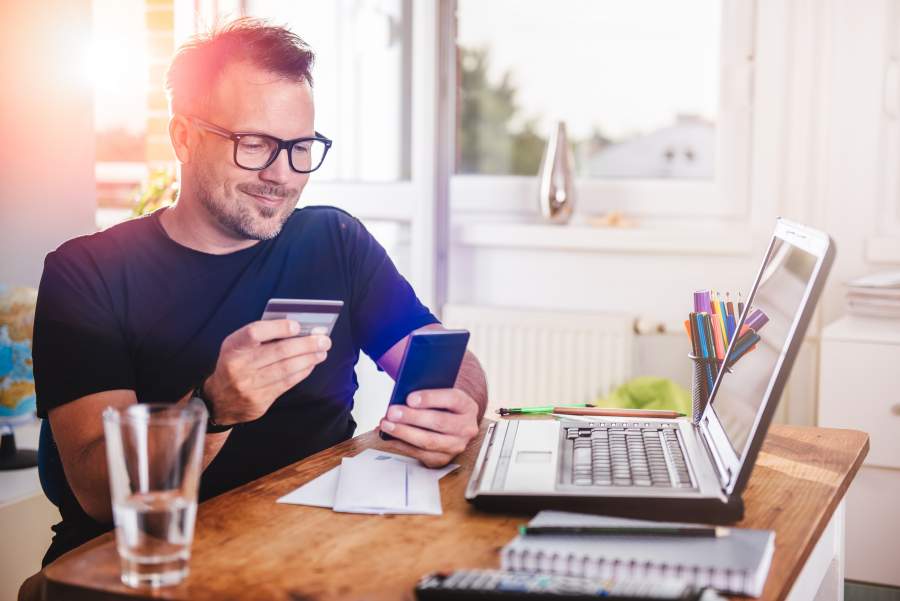 Mann sitzt lächelnd mit Smartphone und Kreditkarte in der Hand vor seinem Laptop am Schreibtisch.