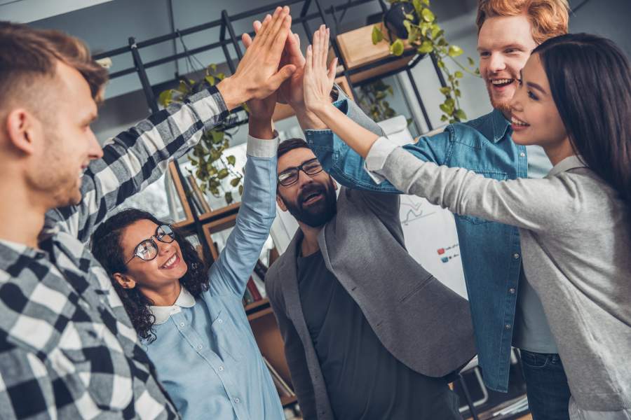 Fünf junge Mitarbeiter und Mitarbeiterinnen stehen zusammen in einem modernen Büro und geben sich ein gemeinsames High-Five.