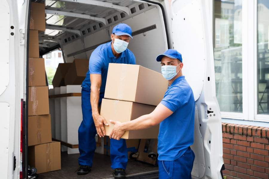 Zwei blau angezogene Mitarbeiter eines Umzugsunternehmens mit Mund-Nasen-Schutz laden Kartons aus einem Transporter aus.