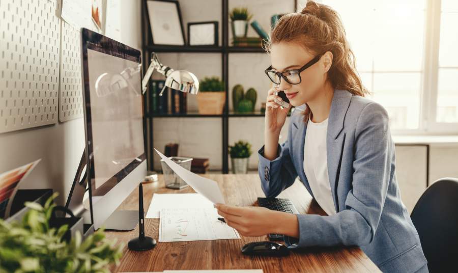 Junge Frau mit Brille in Business-Outfit an Schreibtisch telefoniert und blickt auf Unterlagen in ihrer Hand