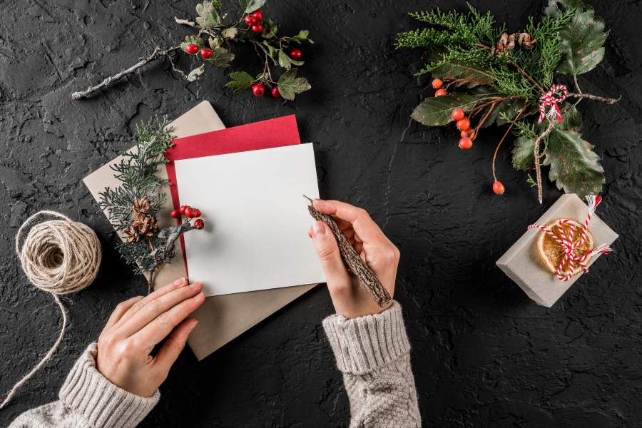 Frau schreibt weihnachtliche Grußkarten während neben ihr verschiedene Bastmaterialien wie Paketschnur, Tannenzweige und getrocknete Beeren liegen.