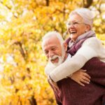 Älteres, lachendes Paare, das im goldenen Herbst draußen im Freien ist, während der Mann seine Frau Huckepack nimmt.
