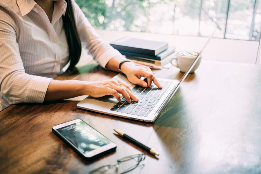 Junge Frau arbeitet an einem Laptop an einem Holztisch, auf dem Smartphone, Brille, Stift, Cappuccino-Tasse und Bücher zu sehen sind