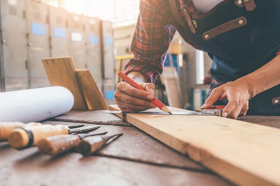 Ein Schreiner in einer Werkstatt zeichnet mit einem Bleistift gerade etwas auf einem Holz an, das auf einer Werkbank neben verschiedenen Werkzeugen liegt.