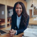 Eine afroamerikanische Geschäftsfrau in Businessoutfit, die mit einem breiten Lachen an einem Schreibtisch sitzt.