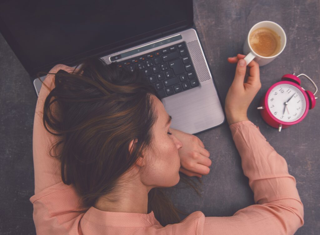Frau in rosa Pulli schläft mit einem Kaffee in der Hand auf ihrem Laptop, neben dem ein analoger Wecker liegt.