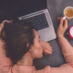 Frau in rosa Pulli schläft mit einem Kaffee in der Hand auf ihrem Laptop, neben dem ein analoger Wecker liegt.