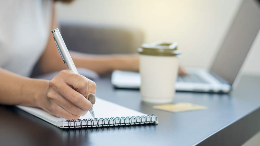 Person arbeitet am Laptop und macht handschriftliche Notizen mit Stift und Block, ein Kaffeebecher steht auf dem Tisch