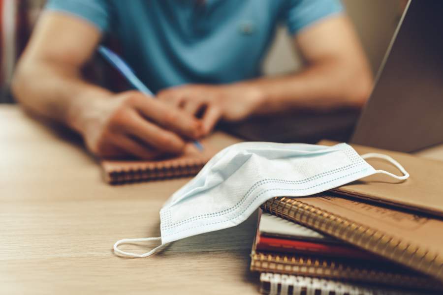 Mann sitzt vor Laptop und macht handschriftliche Notizen auf einem Block, im Vordergrund sind weitere Bläcke und ein Mundnasenschutz zu sehen