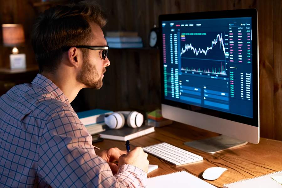 Mann in kariertem Hemd und Brille betrachtet den Kurs einer Crypto-Währung auf einem Monitor und macht sich handschriftliche Notizen