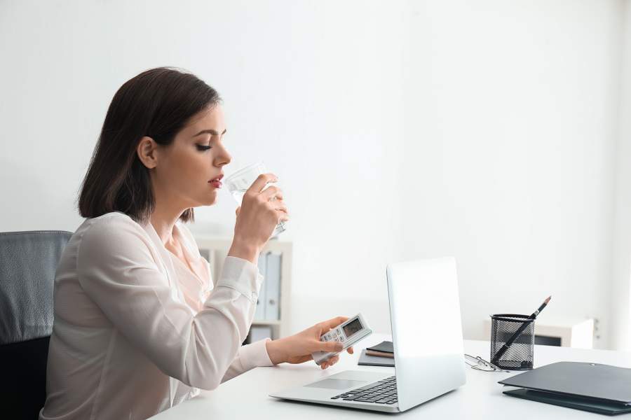 Junge Frau in Büro sitzt an Schreibtisch mit Laptop, Unterlagen und Stiften und trinkt ein Glas Wasser