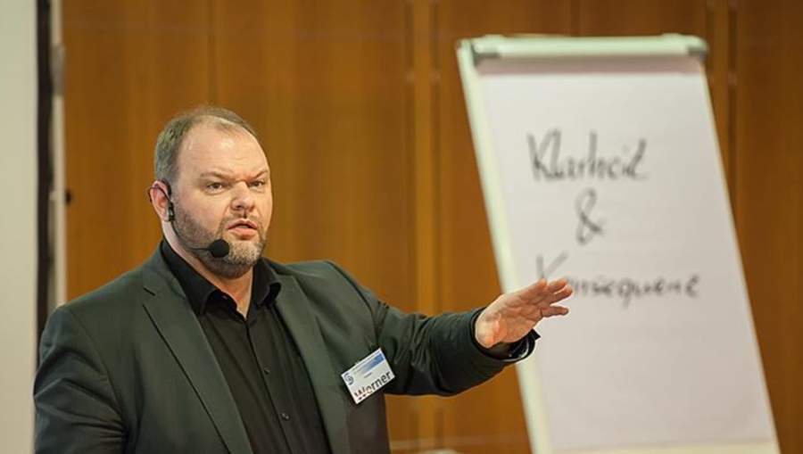 Torsten Werner im Vortrag mit Mikrofon und Namensschild vor einer Flipchart, Schriftzug „Klarheit und Konsequenz“