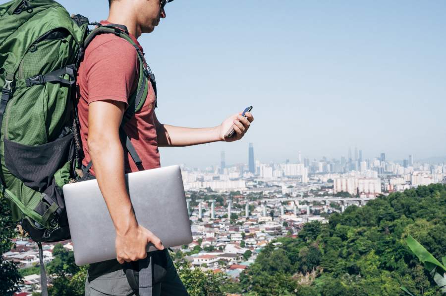 Digitaler Nomade mit rgoßem Rucksack und Laptop blickt aufs Handy, im Hintergrund eine Stadt-Skyline