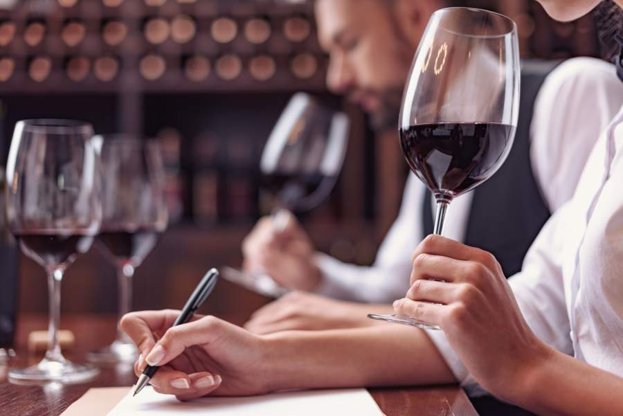 Eine Sommelière beim Weintasting und bei der schriftlichen Bewertung des Rotweins im Glas, im Hintergrund ein Sommelier