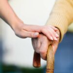 Altenpflegerin legt eine Hand auf die Hand einer alten Person, die eine Gehstock hält