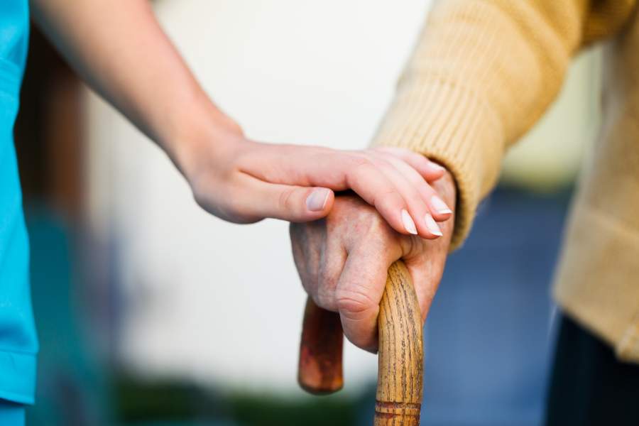 Altenpflegerin legt eine Hand auf die Hand einer alten Person, die eine Gehstock hält