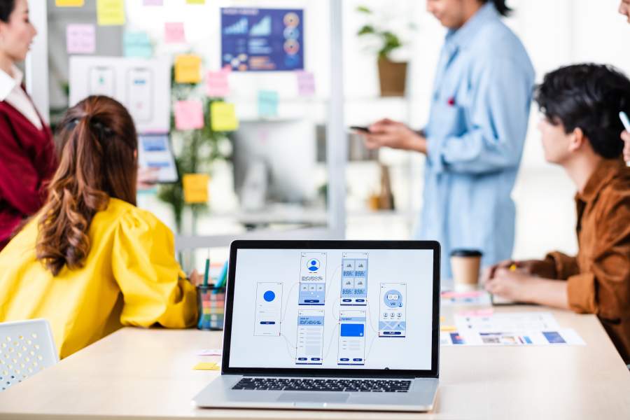 Menschen in einem kreativen Agentur-Arbeitsumfeld in einem Meetingraum, im Vordergrund steht ein Laptop mit Screen-Design