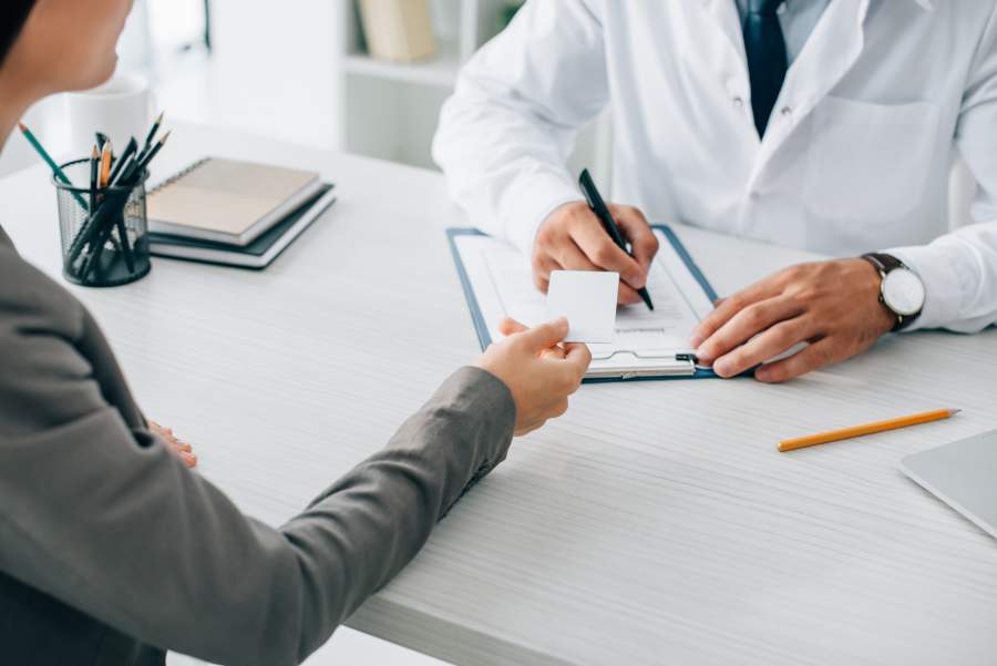 Patientin und Arzt in Sprechstunde, sie reicht ihm einen Zettel, er macht Notizen auf einem Formular