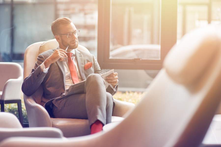 Ein gut gekleideter Geschäftsmann in Anzug sitzt in einer Business-Lounge und sieht von einer Zeitung auf