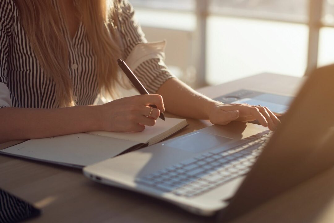 Frau beim Texte schreiben mit Stift in einer Hand und Hand auf Notebook-Tastatur